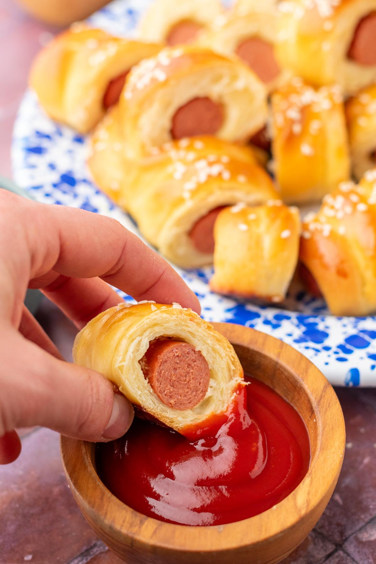 Hand dipping a sourdough pigs in a blanket in ketchup with more on a plate behind.