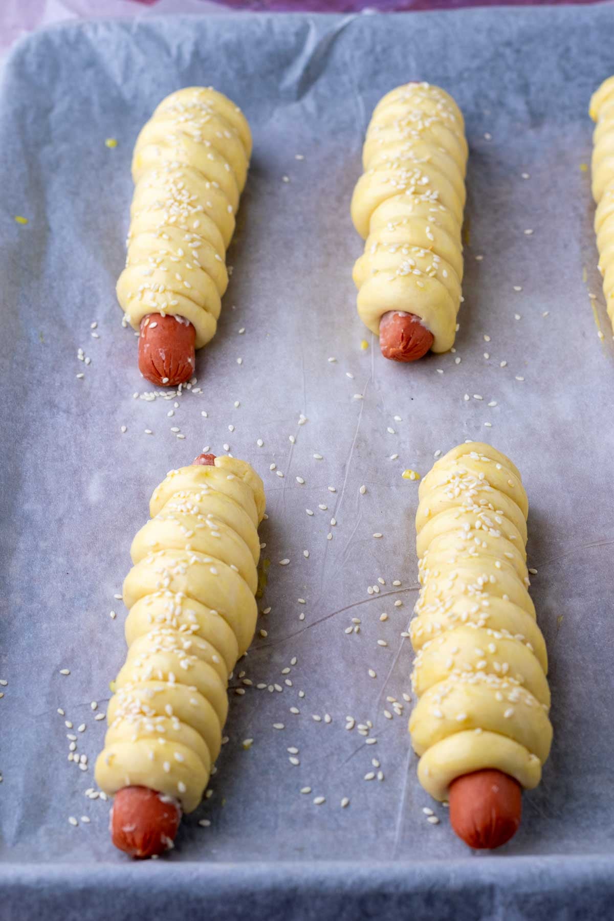 Four sourdough pigs in a blanket on a baking sheet at the end of final proof with an egg wash and sesame seeds.