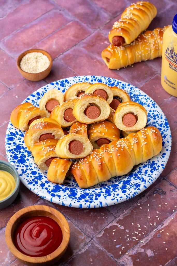 Sourdough pigs in a blanket on a blue and white enamel plate surrounded by sesame seeds, mustard, and ketchup.