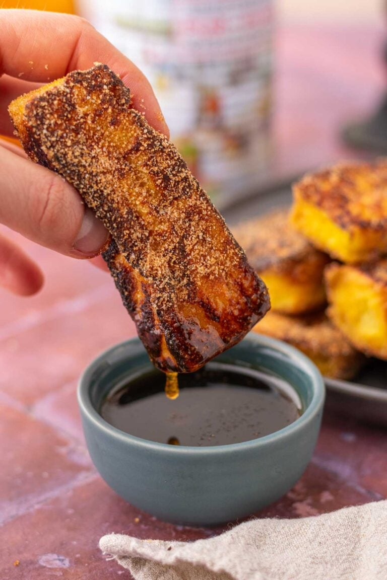 Hand dipping a sourdough cinnamon french toast stick in maple syrup with syrup dripping.