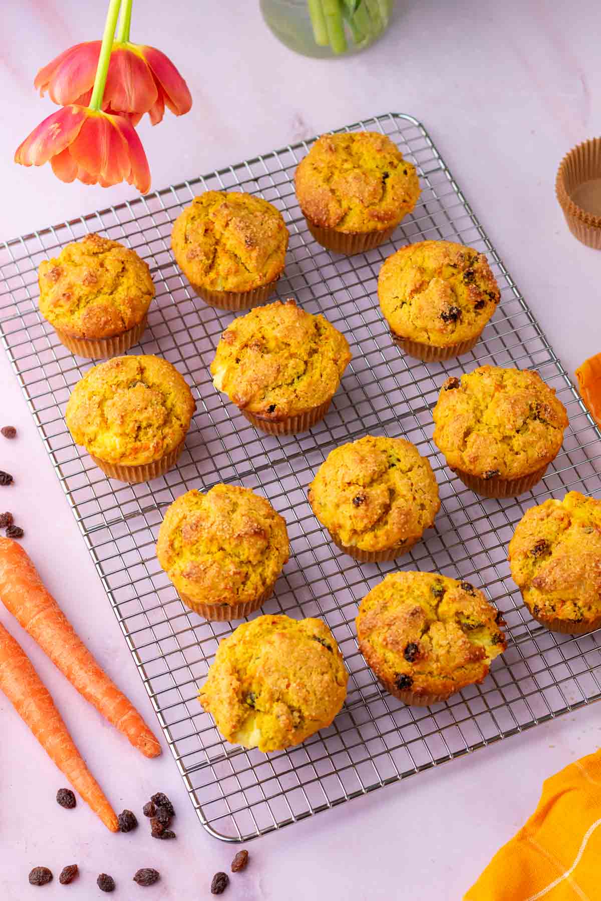 Twelve sourdough carrot cake muffins on a wire rack surrounded by carrots and raisins.
