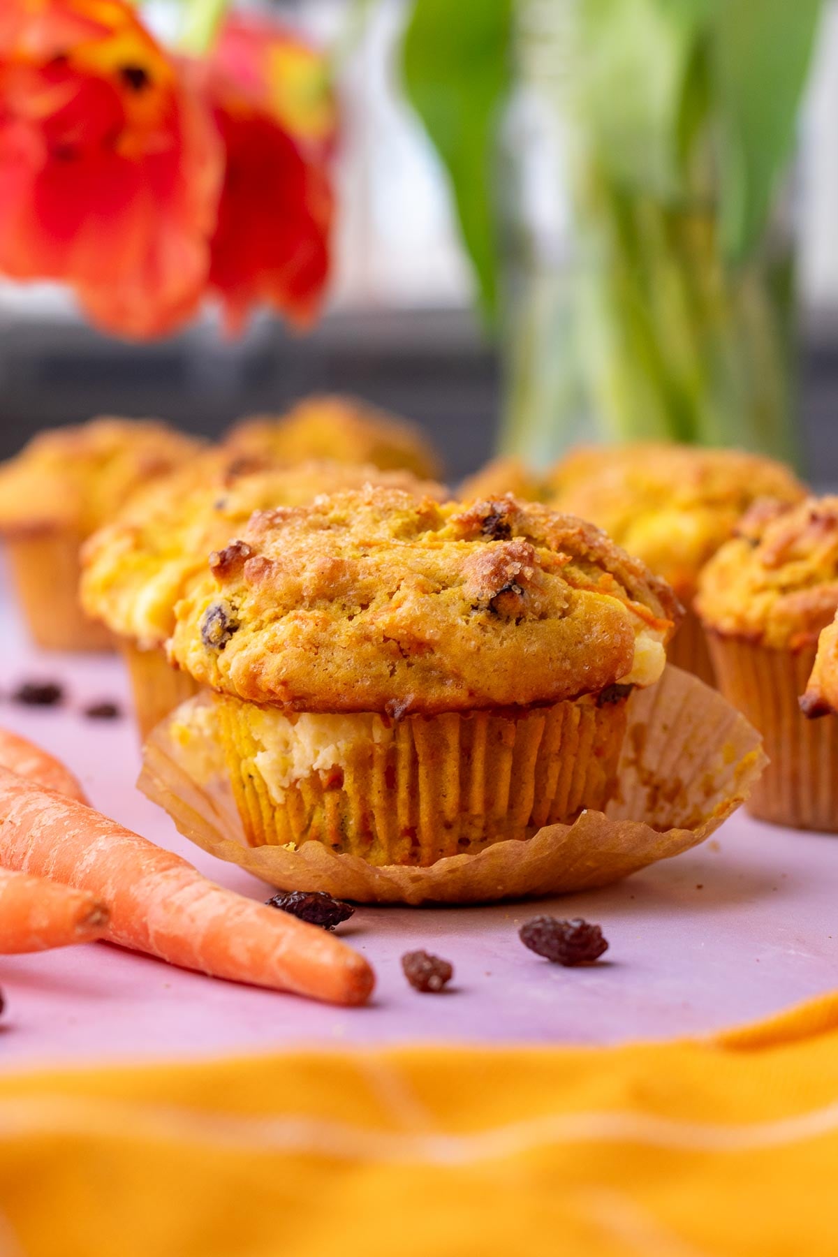 Sourdough carrot cake muffins with cream cheese surrounded by carrots, raisins, and tulips in the background.