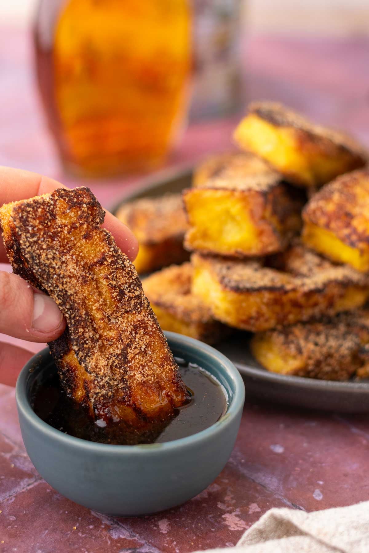 Hand dipping a sourdough cinnamon french toast stick into a small bowl of maple syrup with more sticks in the background.