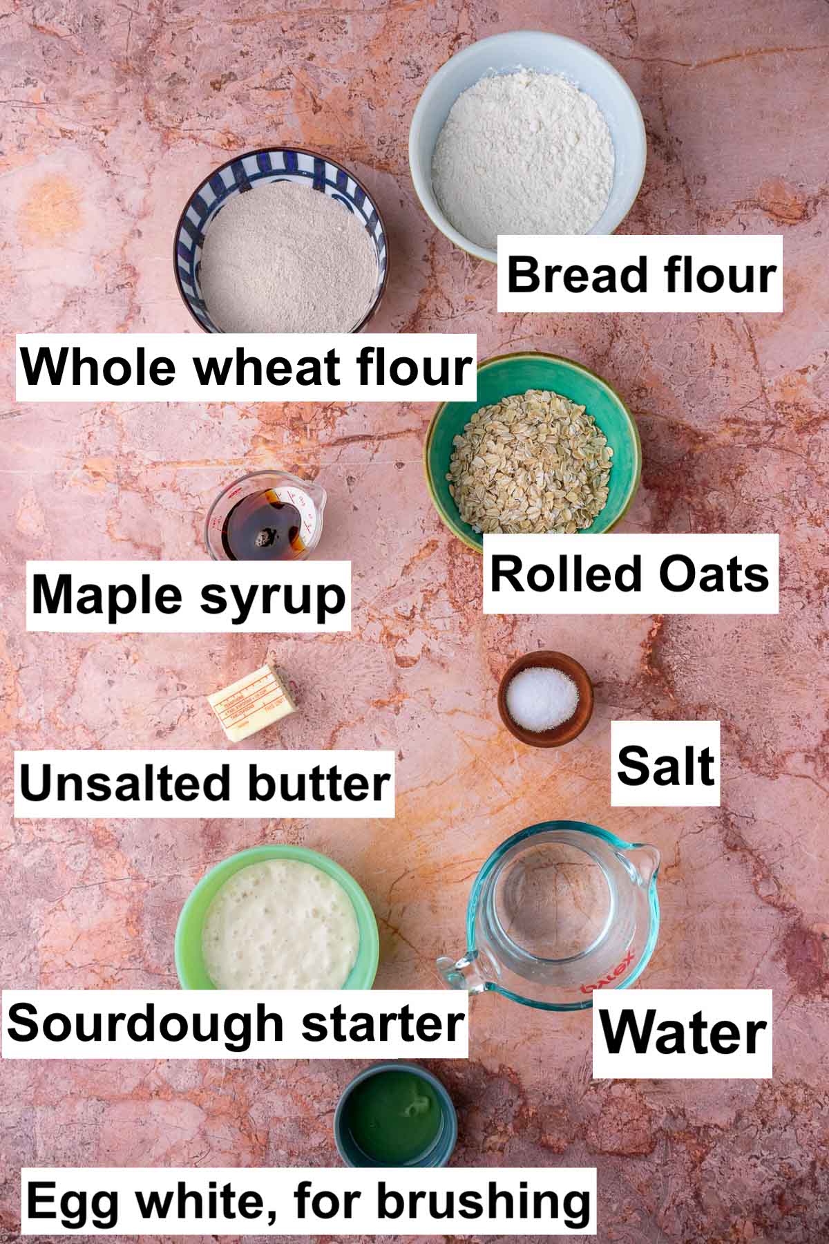 Labeled bowls of ingredients needed for maple oat sourdough sandwich bread including bread flour, whole wheat flour, rolled oats, maple syrup, salt, unsalted butter, water, sourdough starter, and egg white.