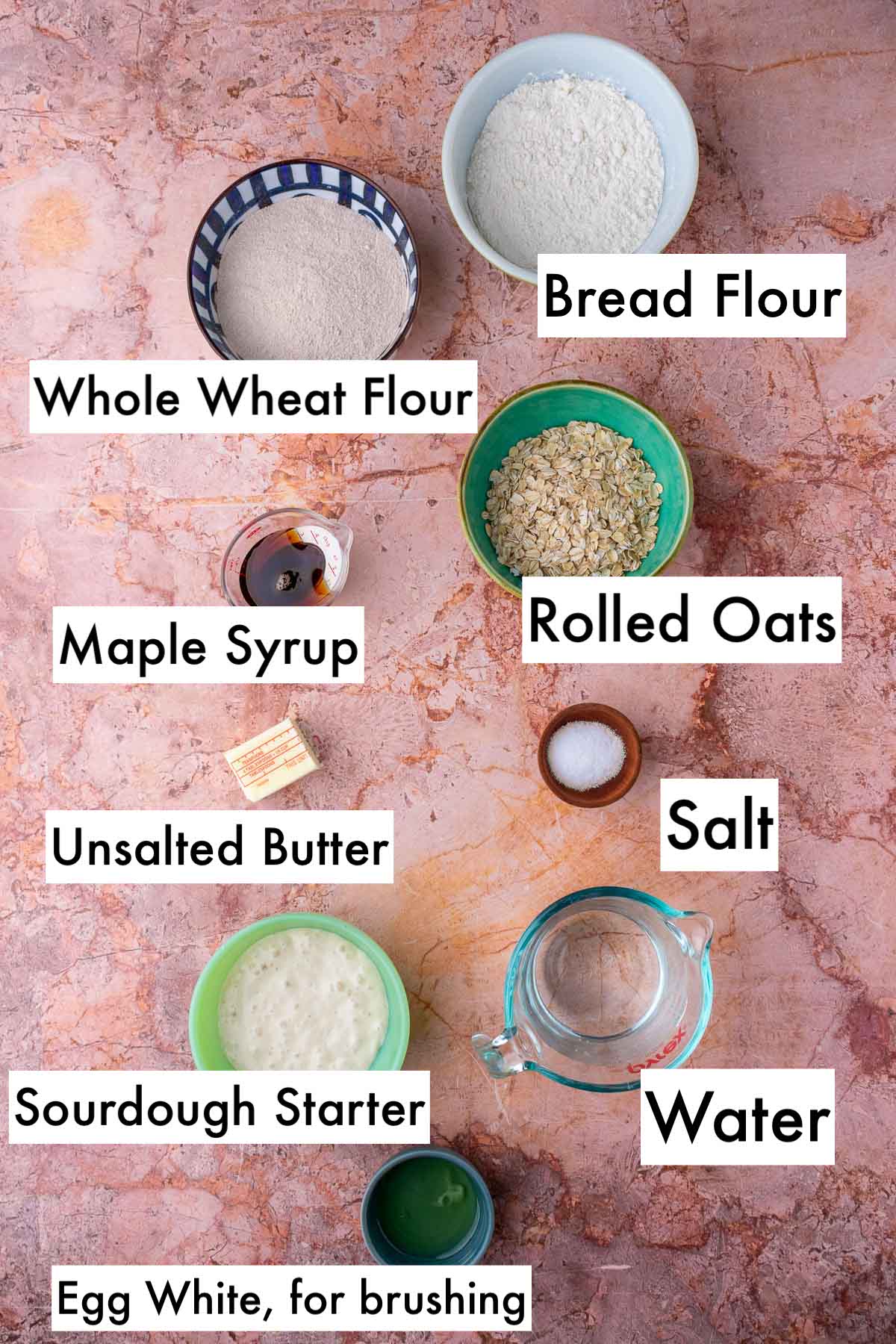 Labeled bowls of ingredients needed for maple oat sourdough sandwich bread including bread flour, whole wheat flour, rolled oats, maple syrup, salt, unsalted butter, water, sourdough starter, and egg white.