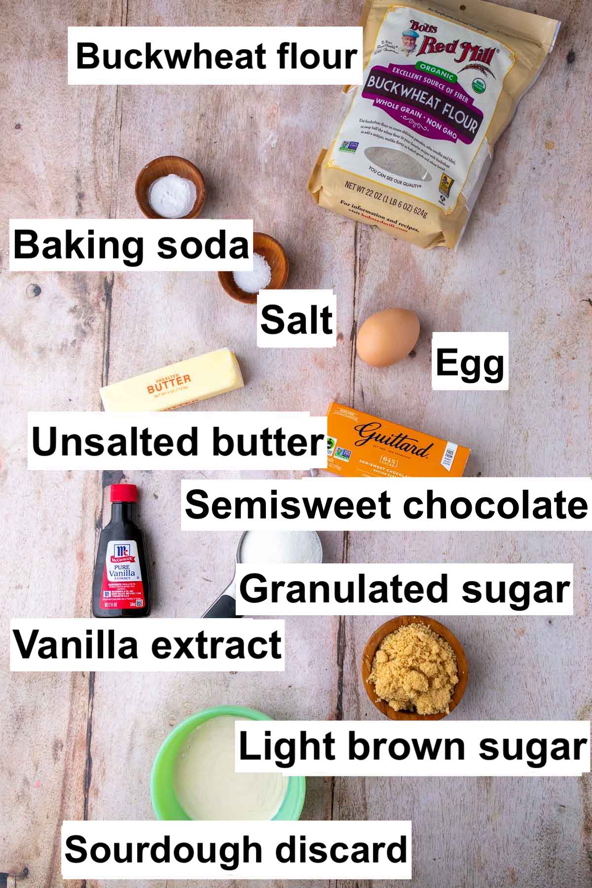 Labeled ingredients needed for sourdough buckwheat chocolate chip cookies including buckwheat flour, baking soda, salt, egg, unsalted butter, semisweet chocolate, vanilla extract, granulated sugar, light brown sugar, and sourdough discard.