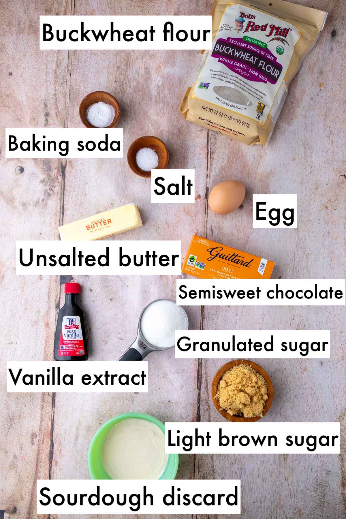 Labeled ingredients needed for sourdough buckwheat chocolate chip cookies including buckwheat flour, baking soda, salt, egg, unsalted butter, semisweet chocolate, vanilla extract, granulated sugar, light brown sugar, and sourdough discard.
