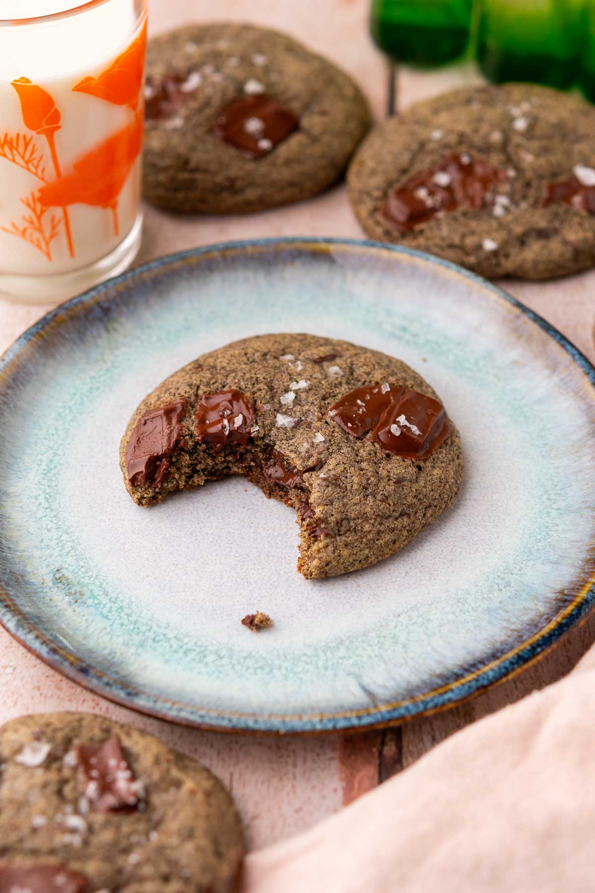 A bite taken out of a sourdough buckwheat chocolate chip cookie on a plate with a glass of milk and more cookies behind it.