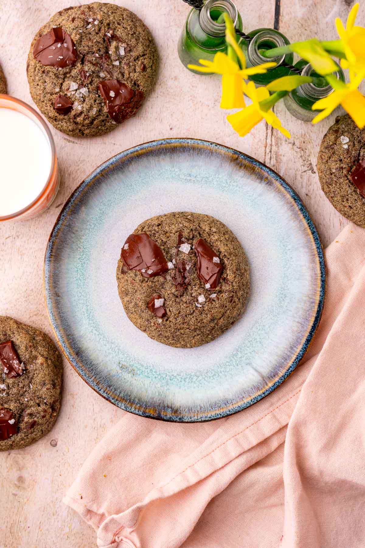 Sourdough buckwheat chocolate chip cookies on a plate, a pink towel, a glass of milk, and daffodils.