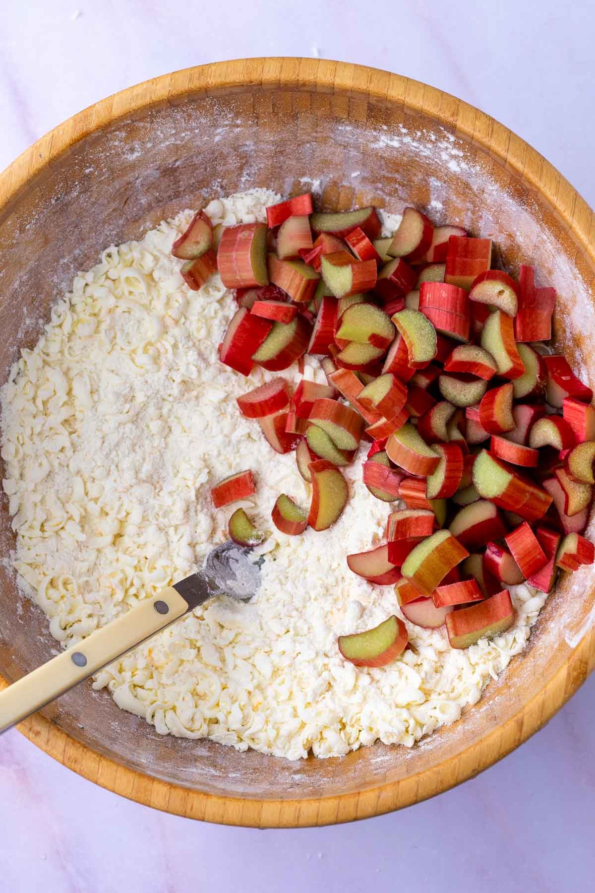 Chopped rhubarb added to a large wooden bowl of dry ingredients for sourdough rhubarb scones with a fork.