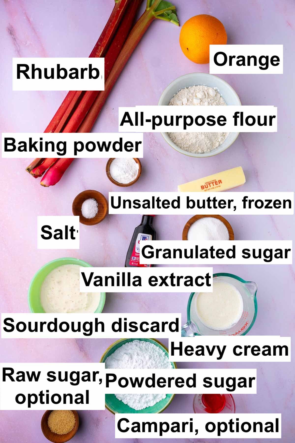 Labeled bowls of ingredients for sourdough rhubarb scones including rhubarb stalks, an orange, all-purpose flour, baking powder, unsalted butter, salt, vanilla, granulated sugar, sourdough discard, heavy cream, powdered sugar, raw sugar, and Campari.