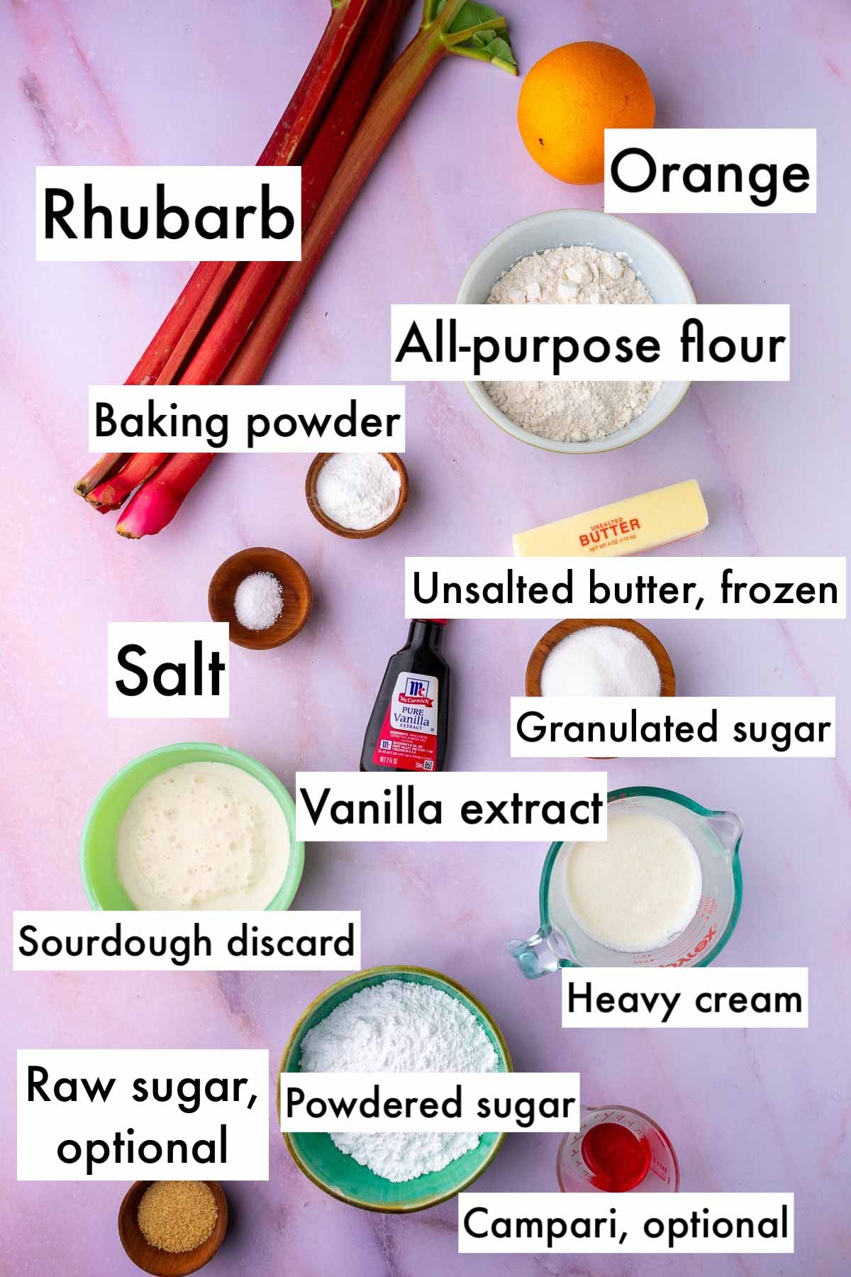 Labeled bowls of ingredients for sourdough rhubarb scones including rhubarb stalks, an orange, all-purpose flour, baking powder, unsalted butter, salt, vanilla, granulated sugar, sourdough discard, heavy cream, powdered sugar, raw sugar, and Campari.