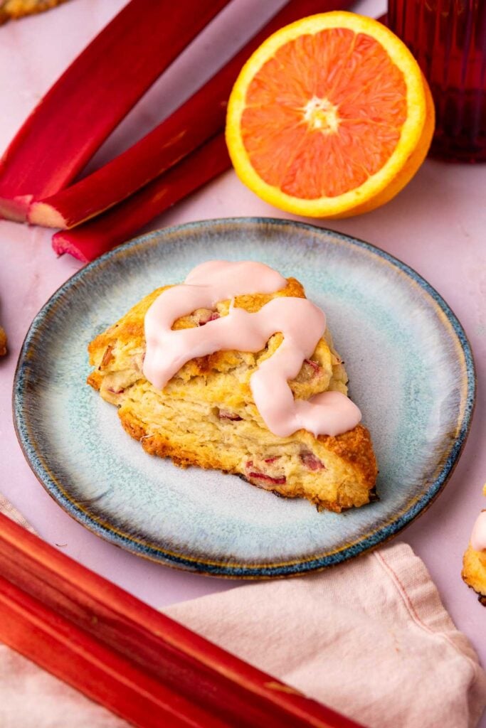Plate with a sourdough rhubarb scone and orange glaze surrounded by red rhubarb stalks and a sliced orange.