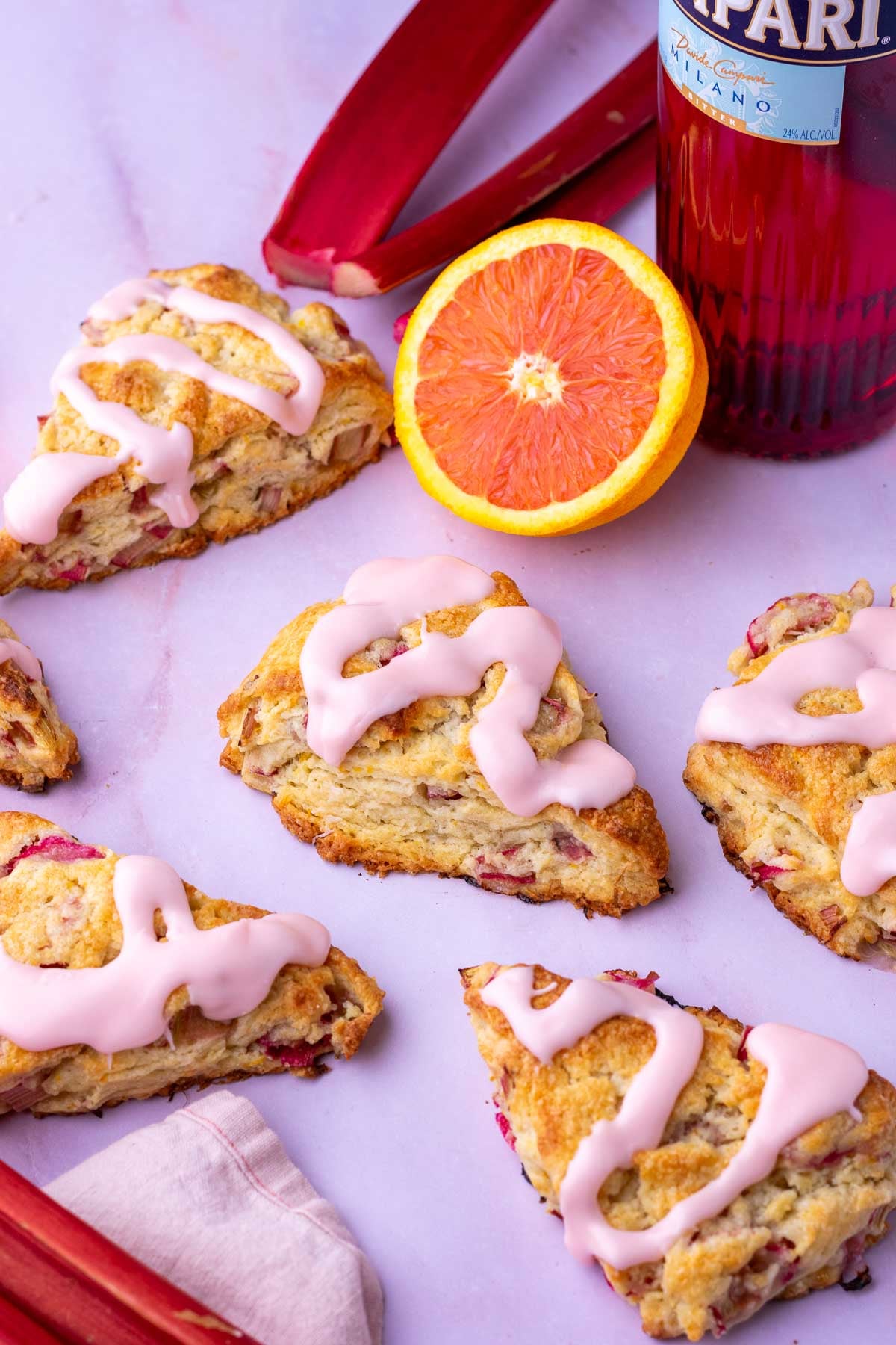 Multiple sourdough rhubarb scones with orange campari glaze surrounded by a sliced orange, bottle of Campari, and rhubarb stalks.