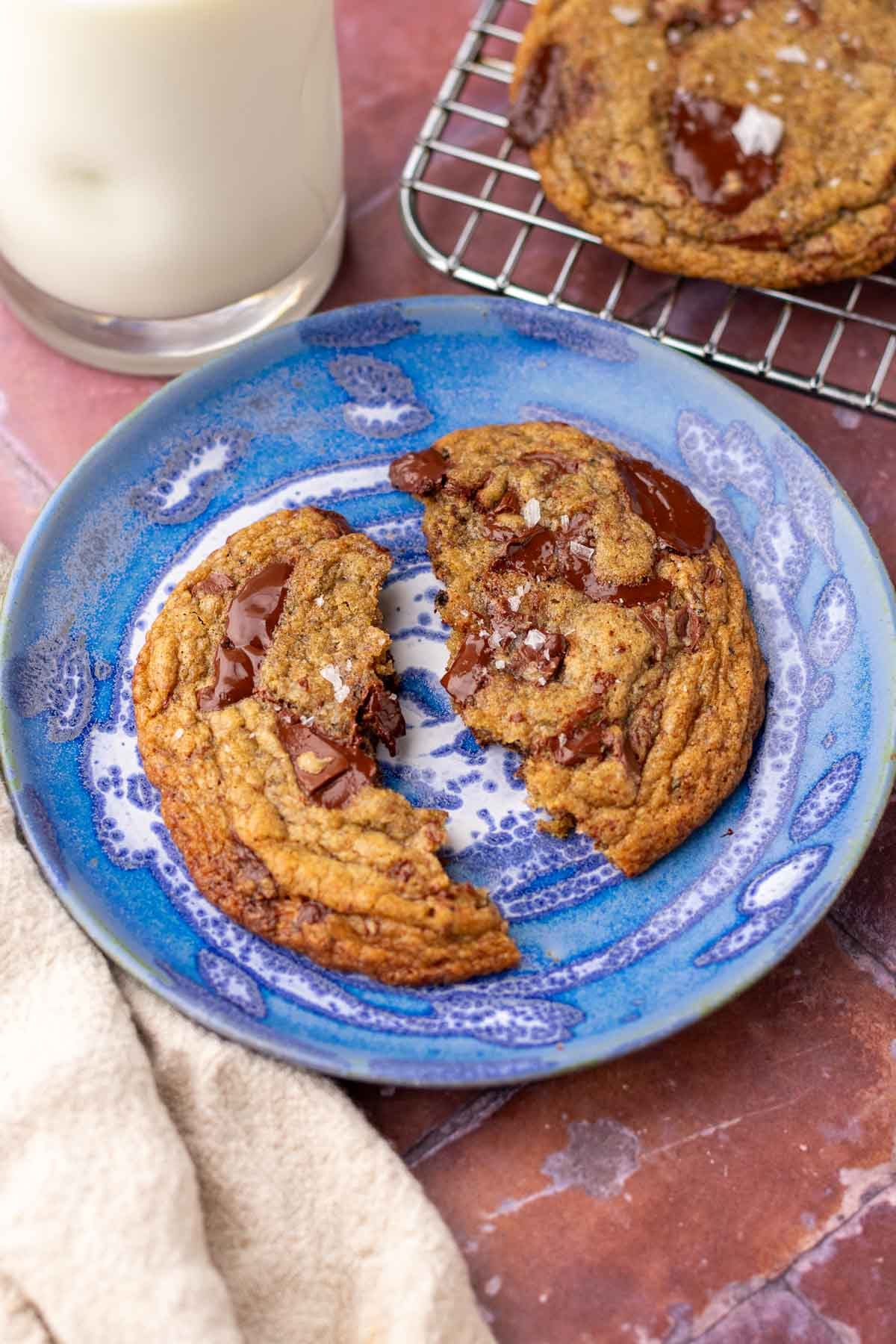 sourdough rye chocolate chip cookie broken in half on a plate with more cookies in the background and a glass of milk.