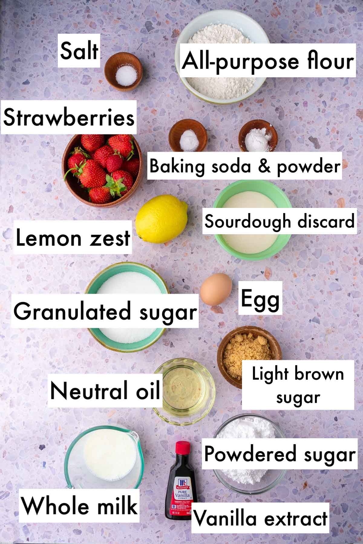 Labeled bowls of ingredients needed for Sourdough Strawberry Bread including all-purpose flour, salt, strawberries, baking soda and baking powder, granulated sugar, lemon zest, egg, brown sugar, oil, milk, powdered sugar, and vanilla extract.