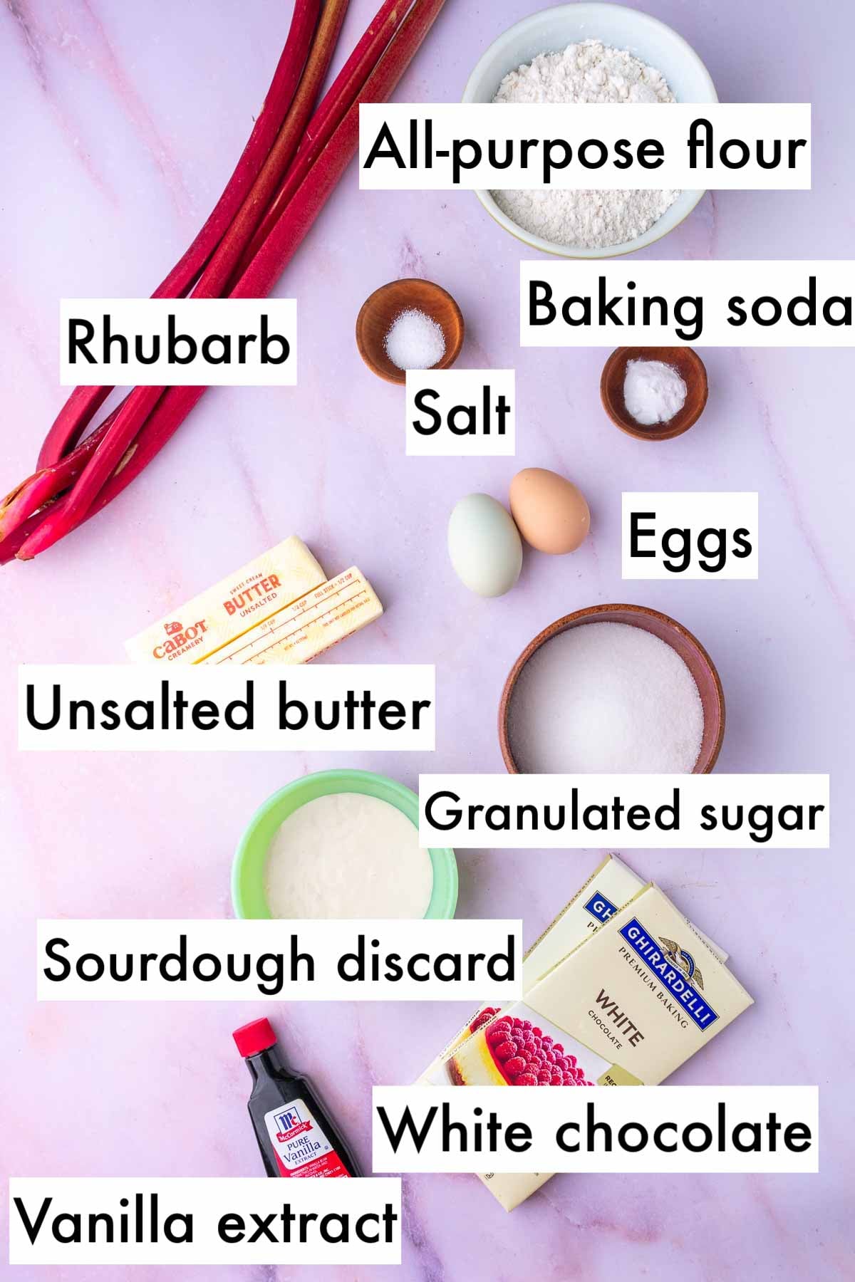 Labeled ingredients needed for sourdough rhubarb cookies including all-purpose flour, baking soda, rhubarb, salt, eggs, unsalted butter, granulated sugar, sourdough discard, white chocolate, and vanilla extract.