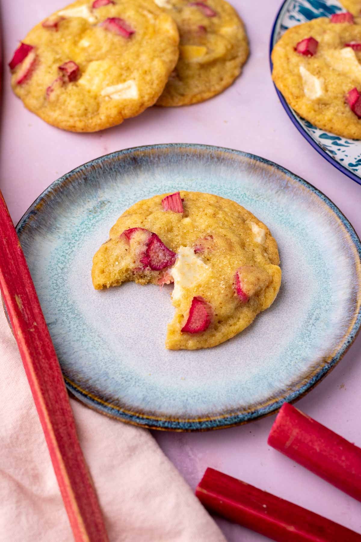 Bite taken out of a sourdough rhubarb cookie on a plate surrounded by more cookies, rhubarb stalks, and a pink towel.