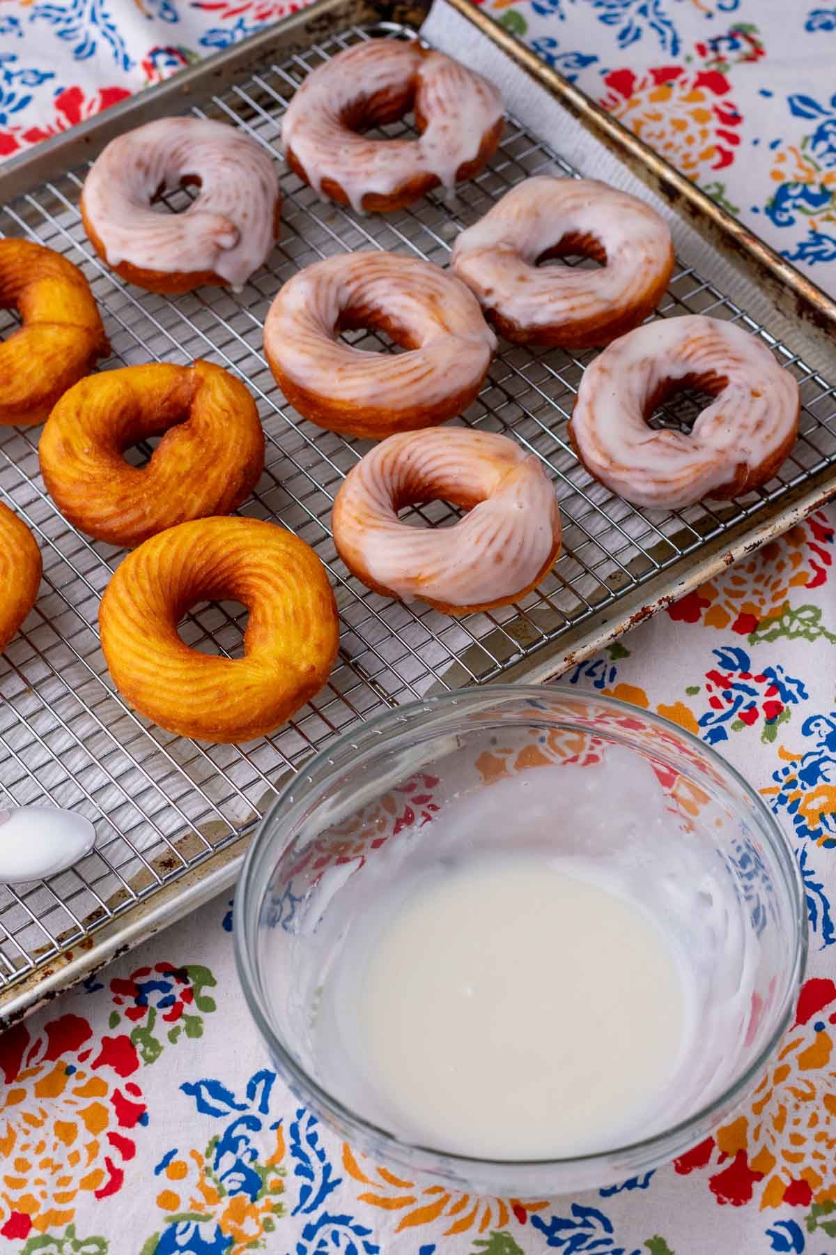 Fried sourdough crullers on a sheet pan with a wire rack and a bowl of glaze in front of the pan.