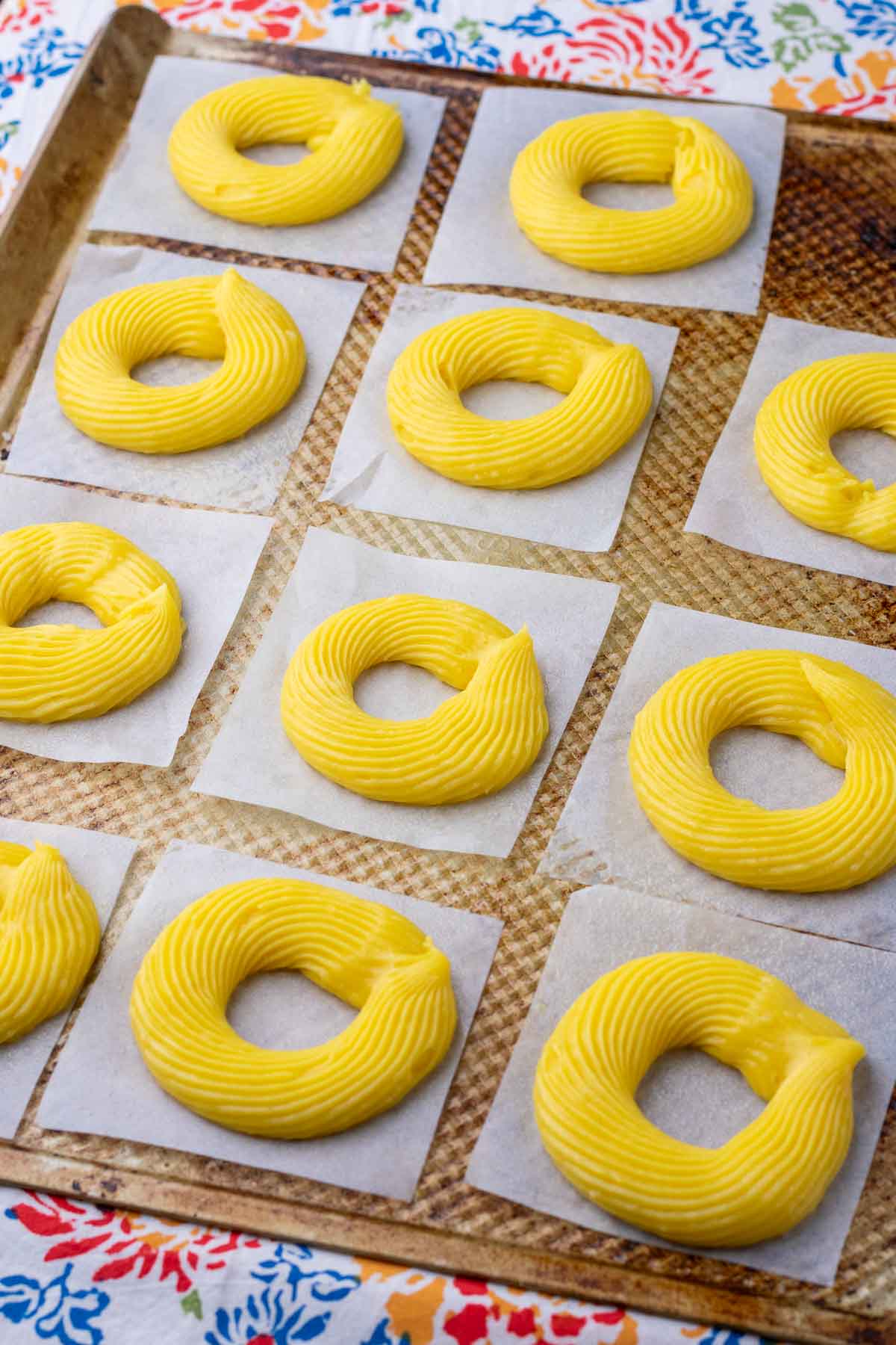 Piped sourdough crullers on a baking sheet with squares of parchment paper.
