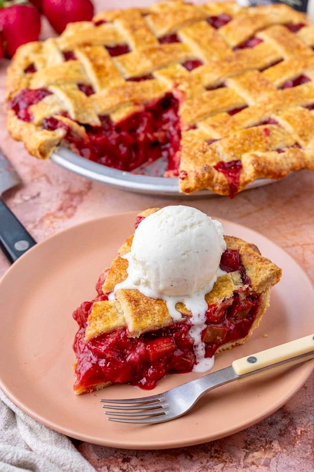 Slice of sourdough strawberry rhubarb pie with a scoop of vanilla ice cream on a plate with a fork and the entire pie in the background.