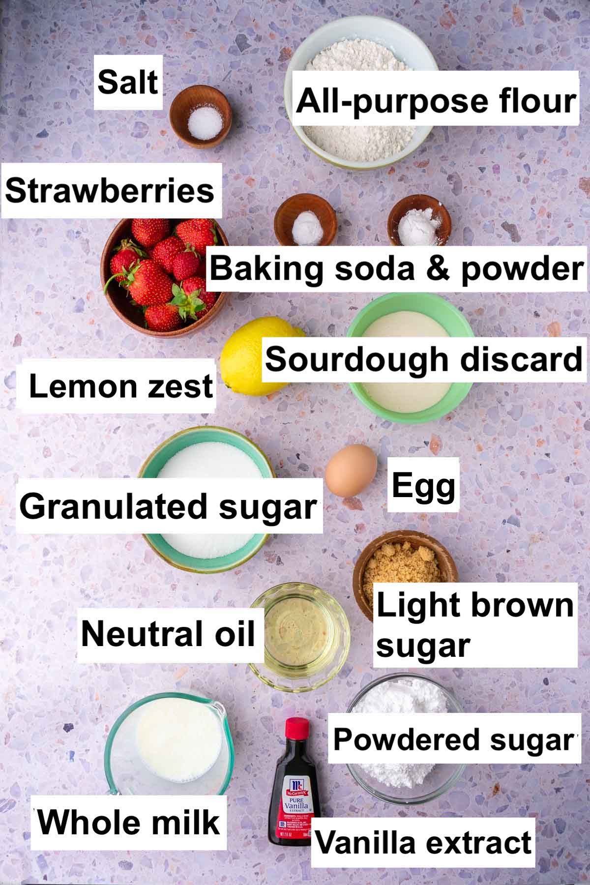 Labeled bowls of ingredients needed for Sourdough Strawberry Bread including all-purpose flour, salt, strawberries, baking soda and baking powder, granulated sugar, lemon zest, egg, brown sugar, oil, milk, powdered sugar, and vanilla extract.