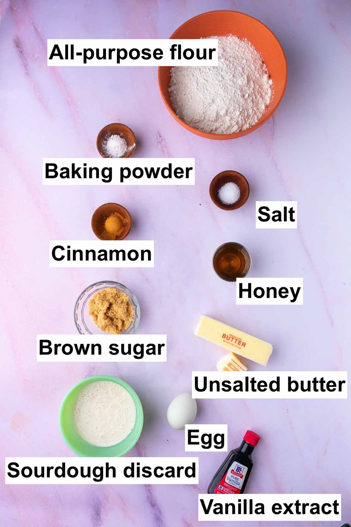 Labeled bowls of ingredients needed for sourdough animal crackers including all-purpose flour, baking powder, salt, cinnamon, honey, brown sugar, unsalted butter, egg, sourdough discard, and vanilla extract.