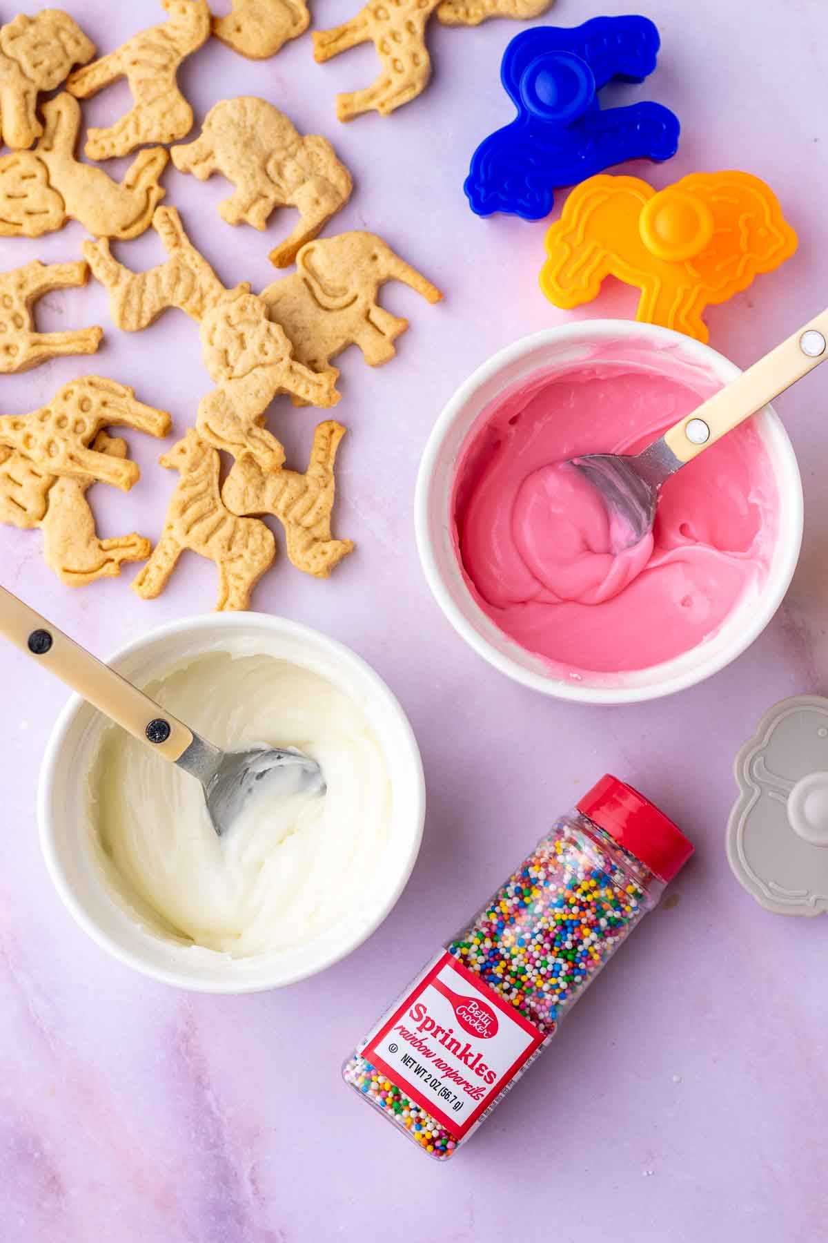 Mixing pink and white frosting for sourdough animal crackers in two bowls with spoons surrounded by crackers and sprinkles.