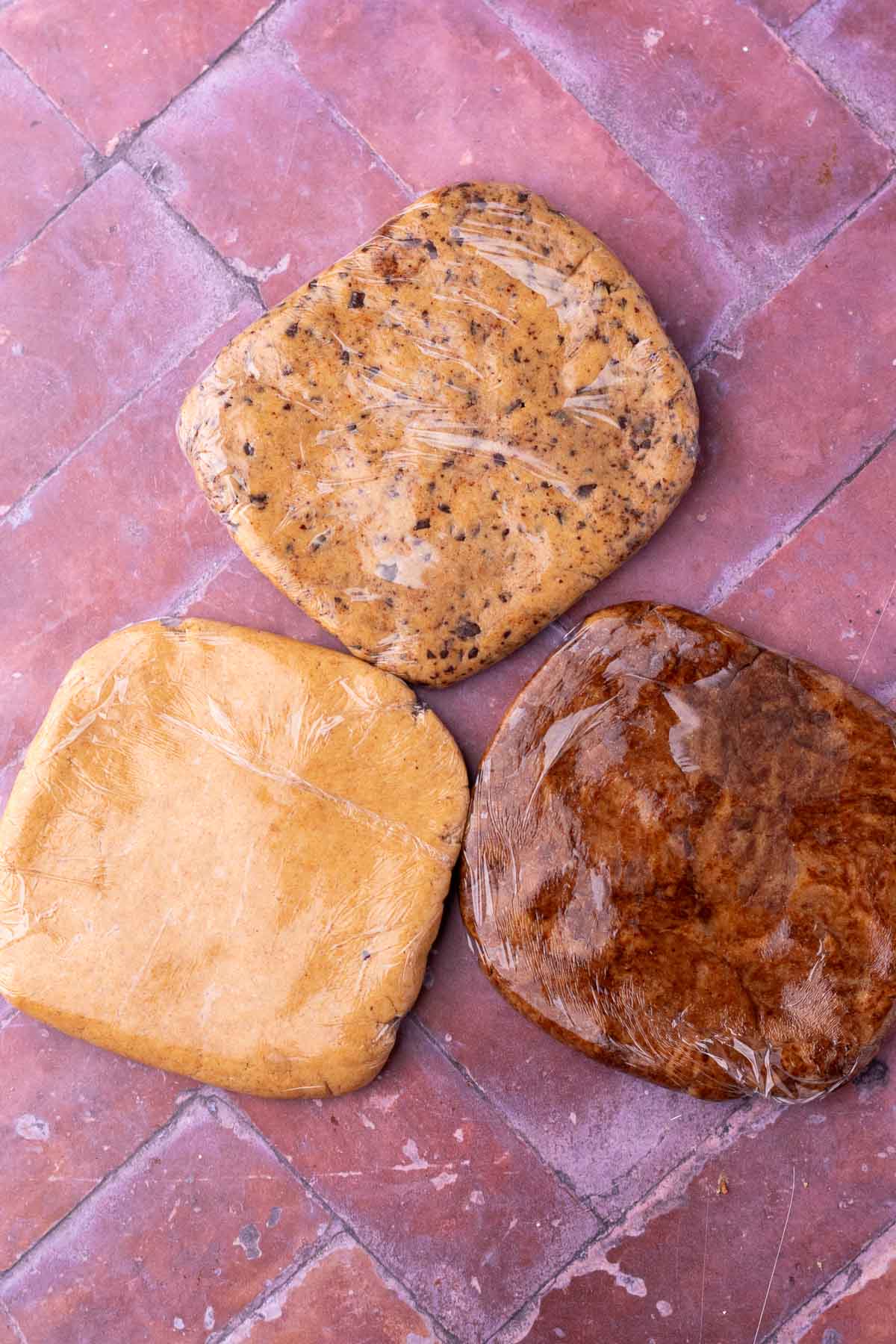 Three flavors of sourdough teddy graham dough wrapped in plastic wrap including cinnamon, chocolate chip, and chocolate.