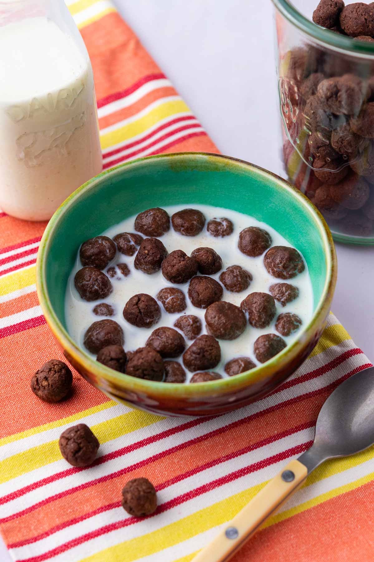 Bowl of sourdough cocoa puffs with milk with a glass of milk and glass jar of cocoa puffs in the background and a spoon in front.