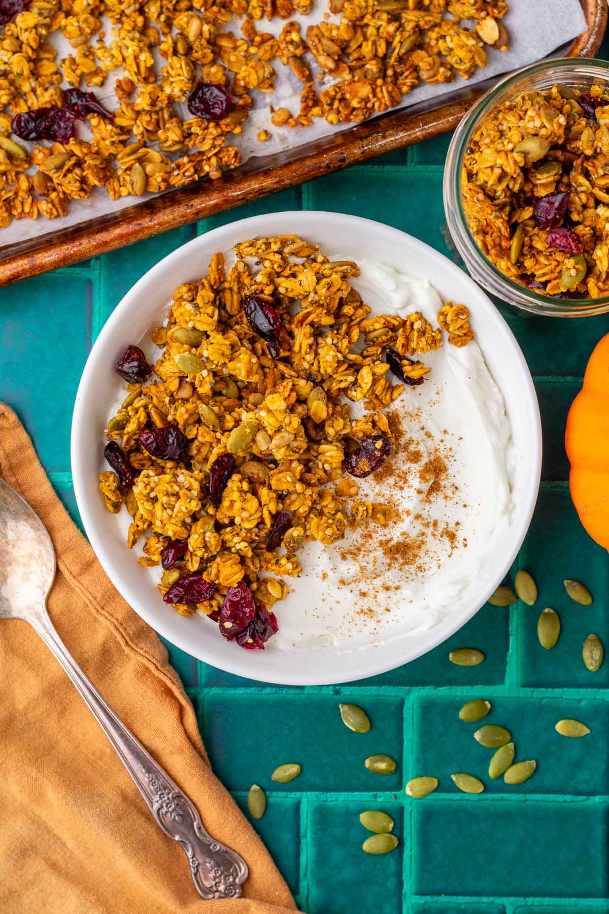 Bowl of yogurt with sourdough pumpkin granola with granola, pumpkin seeds, a spoon and orange towel, and pumpkin surrounding it.