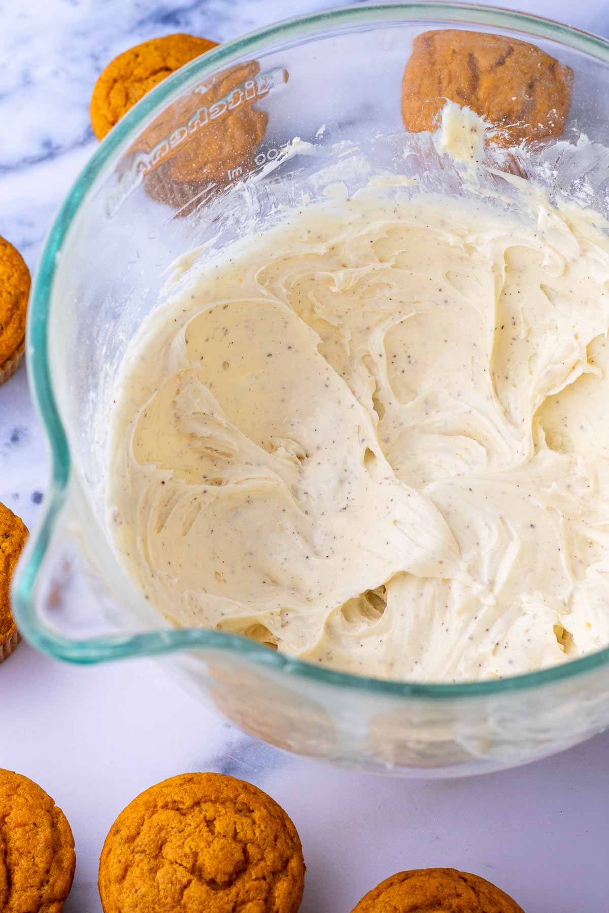 Brown butter cream cheese frosting in a glass stand mixer bowl surrounded by unfrosted sourdough pumpkin cupcakes.