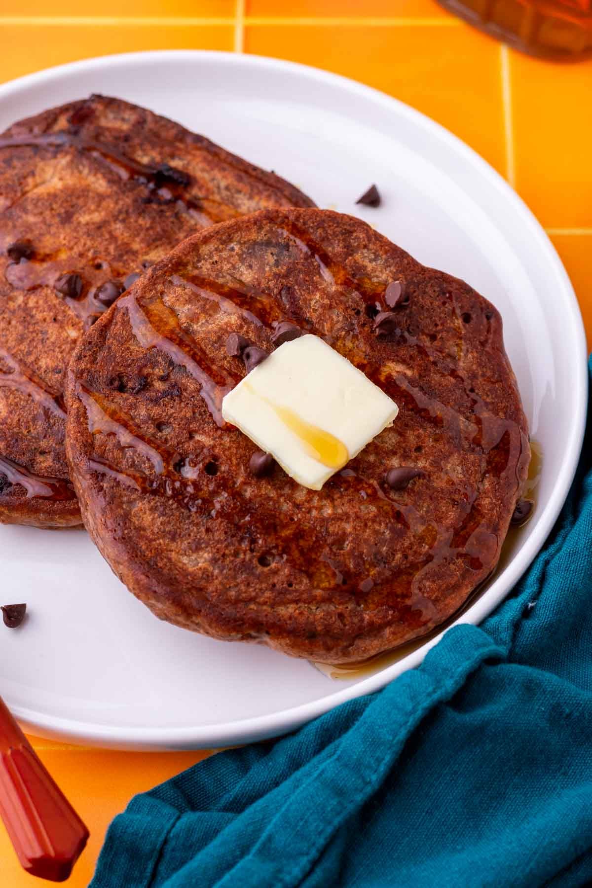 Close up of a sourdough chocolate protein pancake on a plate with maple syrup, chocolate chips, and butter.