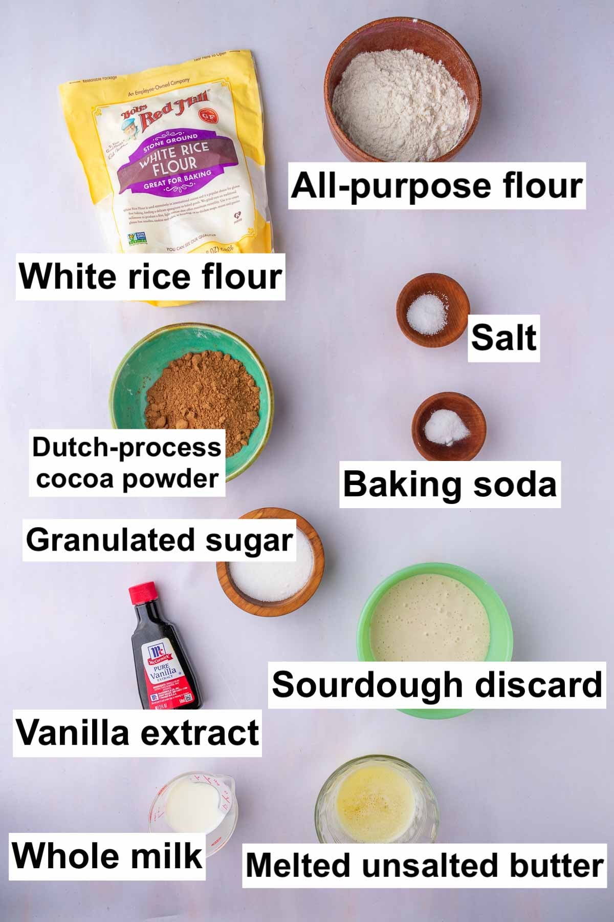 Labeled bowls of ingredients needed for sourdough cocoa puffs including all-purpose flour, white rice flour, salt, baking soda, dutch-process cocoa powder, granulated sugar, sourdough discard, vanilla extract, whole milk, and melted butter.