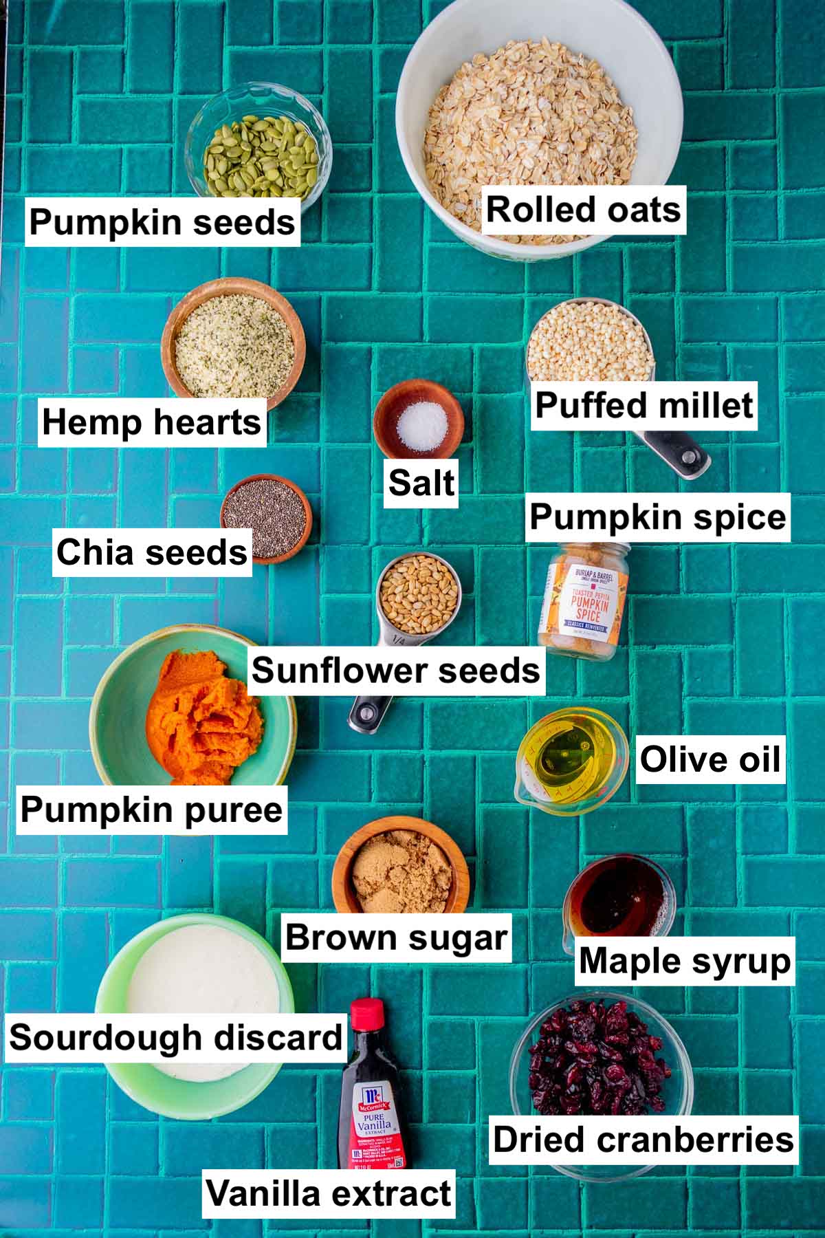 Labeled bowls of ingredients needed for sourdough pumpkin granola including rolled oats, pumpkin seeds, puffed millet, hemp hearts, salt, chia, sunflower seeds, pumpkin spice, olive oil, maple syrup, brown sugar, pumpkin puree, sourdough discard, vanilla extract, and dried cranberries.