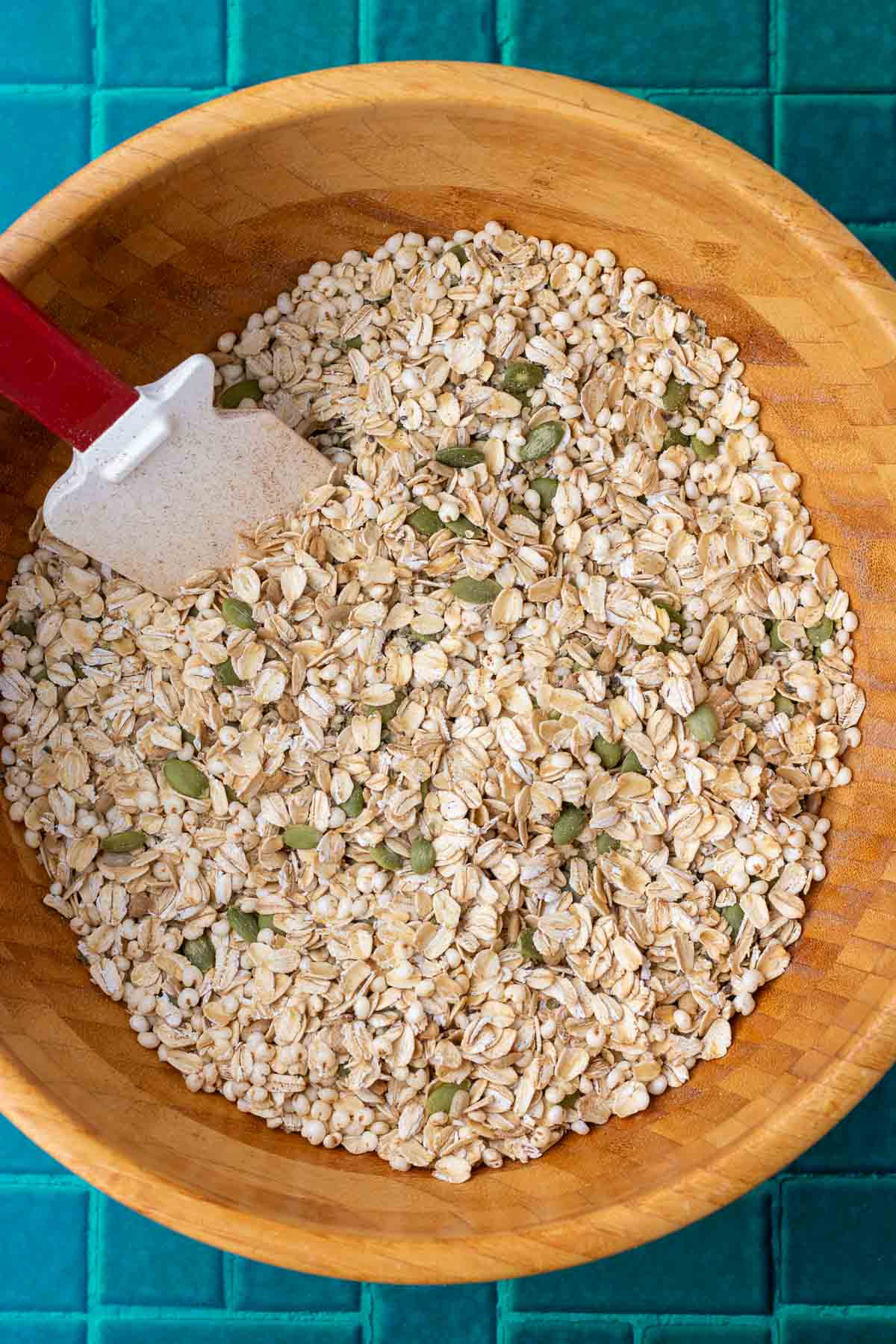 Spatula mixing the dry ingredients for sourdough pumpkin granola in a large wooden bowl including oats, pumpkin seeds, spices, and sunflower seeds.