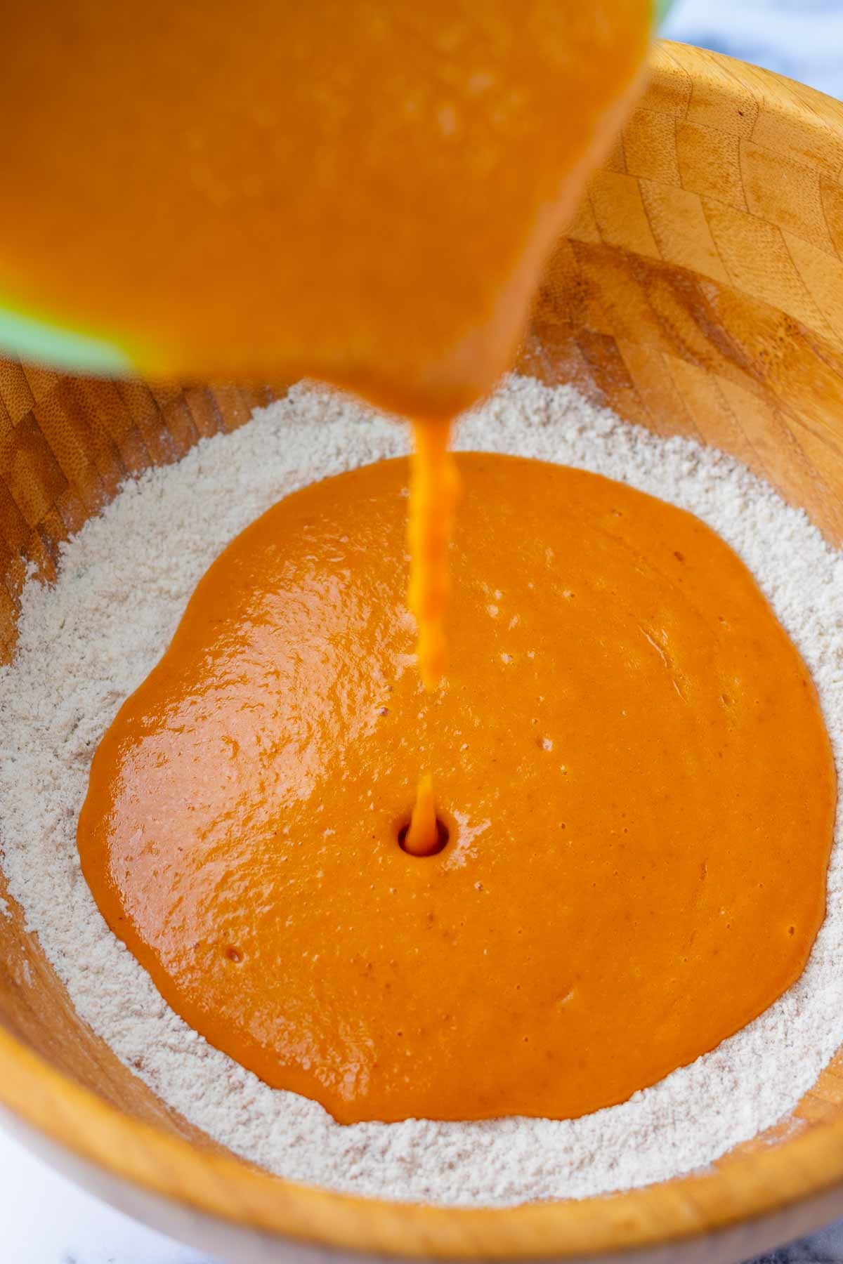 Pouring wet ingredients into a wooden bowl of dry ingredients for sourdough pumpkin cupcakes.