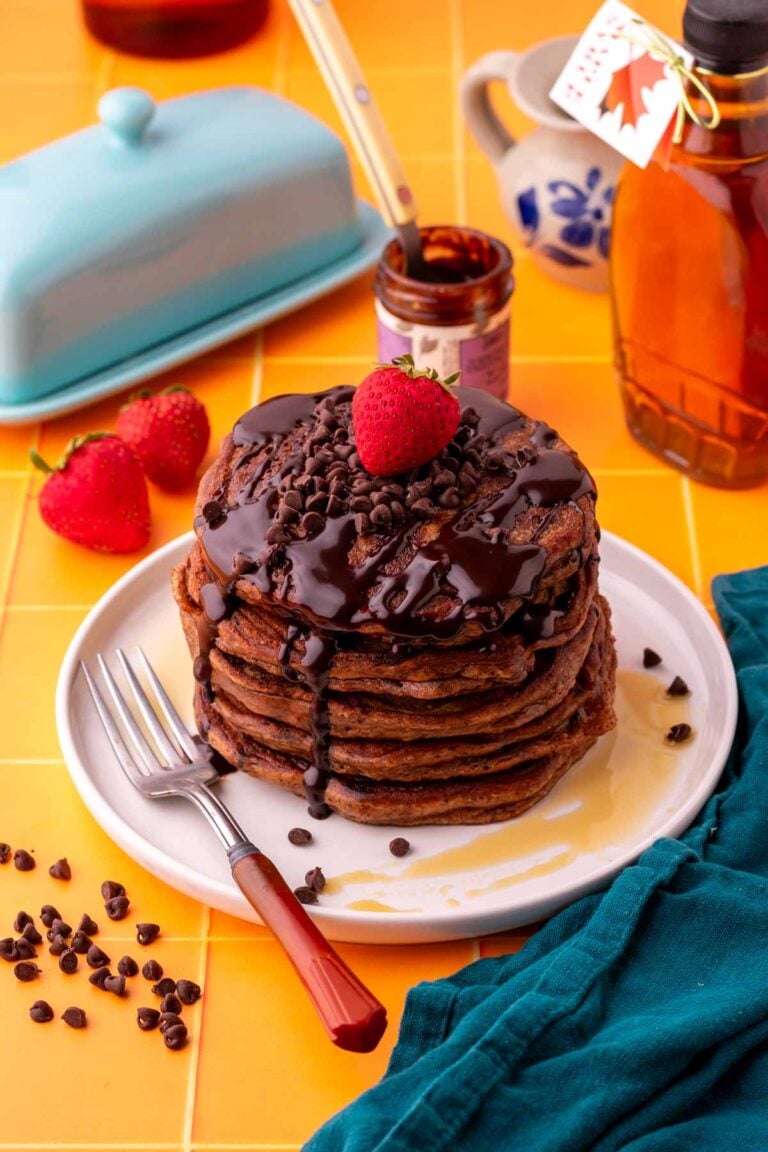 Stack of sourdough chocolate protein pancakes with chocolate syrup, chocolate chips, and a strawberry on top surrounded by a fork, maple syrup, and butter container.
