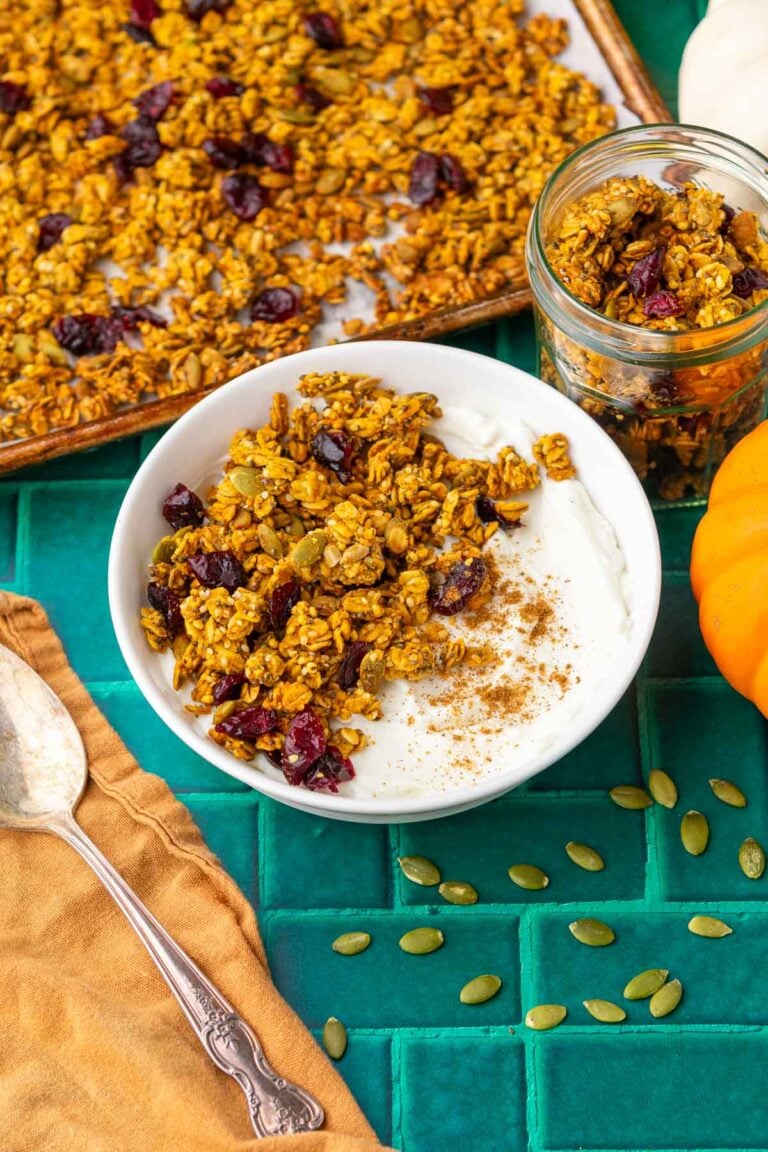 Sourdough pumpkin granola in a bowl with yogurt with more granola surrounding it, pumpkins, and a spoon with a towel.