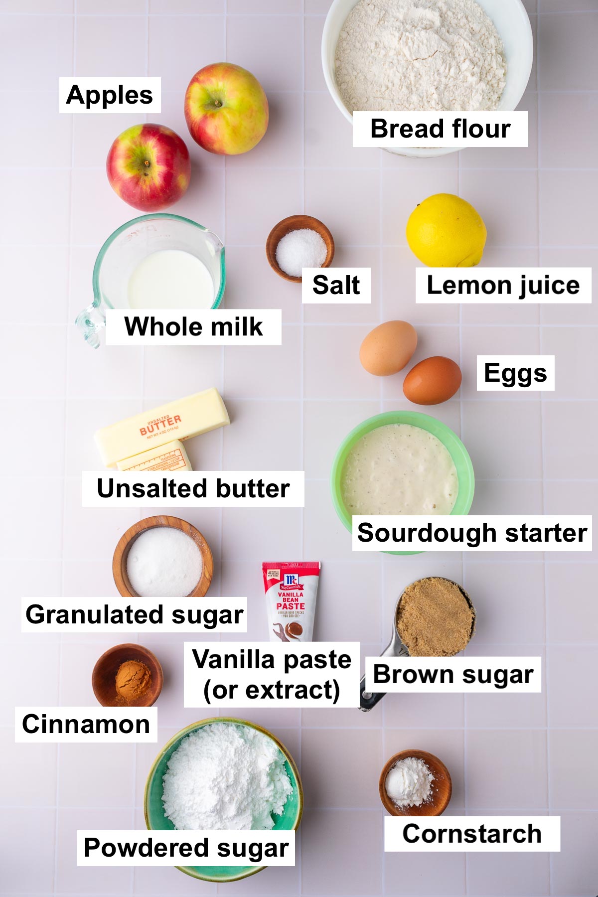 Labels of ingredients needed for sourdough apple cinnamon rolls including apples, bread flour, lemon juice, salt, eggs, whole milk, unsalted butter, sourdough starter, granulated sugar, vanilla, brown sugar, cinnamon, powdered sugar, and cornstarch.