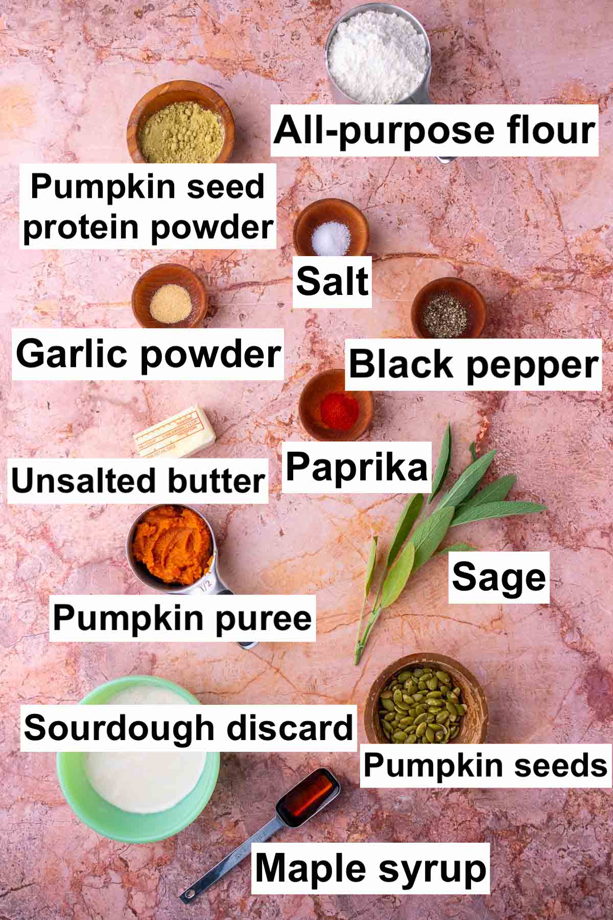 Labeled bowls of ingredients needed for sourdough pumpkin crackers including all-purpose flour, salt, black pepper, garlic powder, paprika, unsalted butter, sage, pumpkin puree, sourdough discard, maple syrup, pumpkin seeds, and pumpkin seed protein powder.