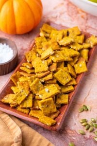 Plate of sourdough pumpkin crackers surrounded by pumpkin seeds and a pumpkin.
