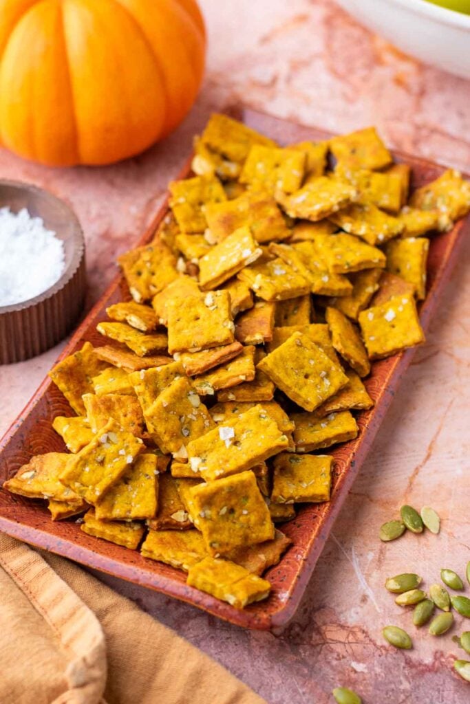 Plate of sourdough pumpkin crackers surrounded by pumpkin seeds and a pumpkin.